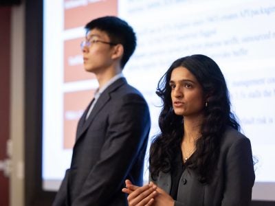 Two students standing in front of a screen and speaking to their classmates, not shown in the photo.