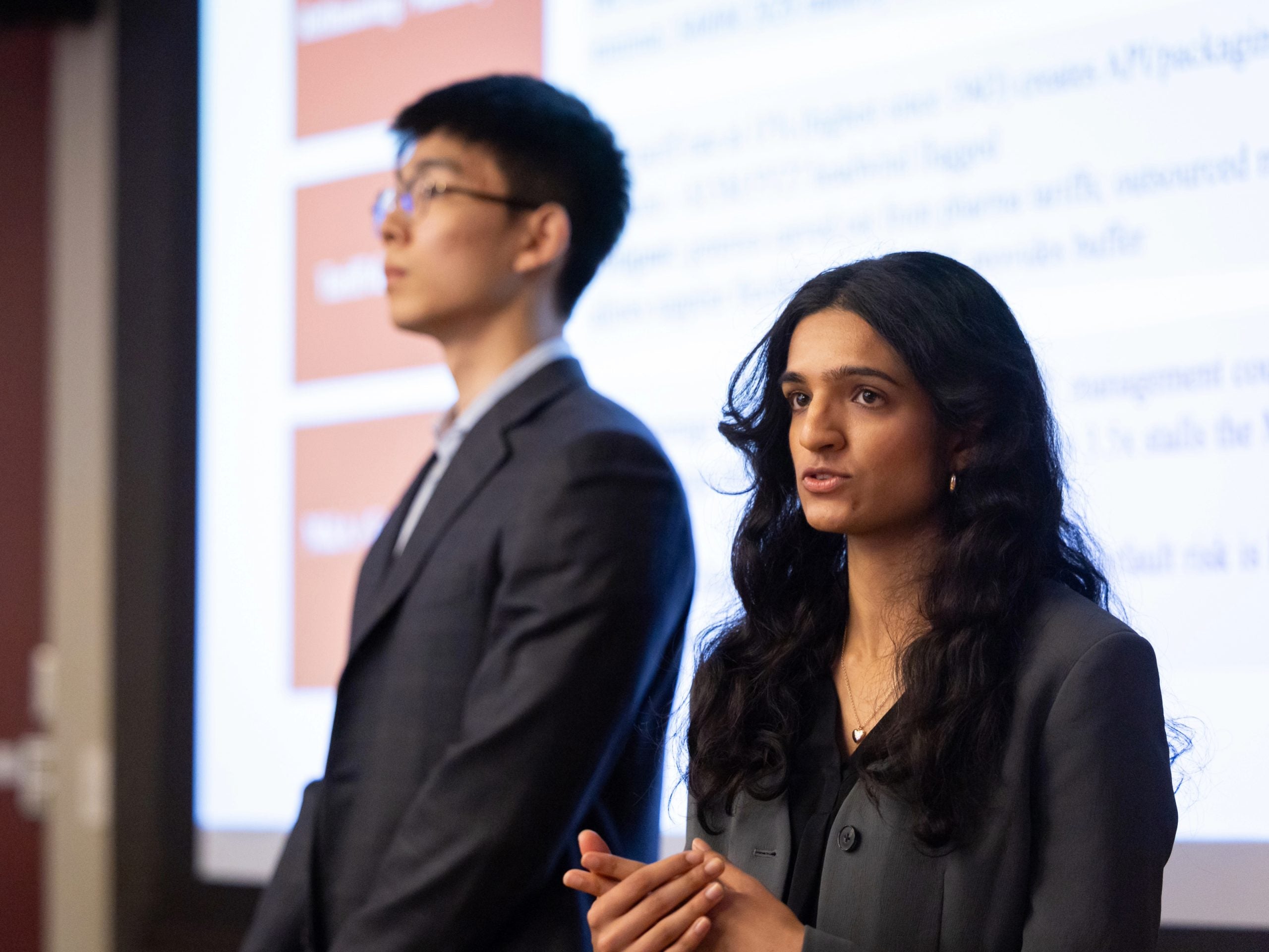 Two students standing in front of a screen and speaking to their classmates, not shown in the photo.