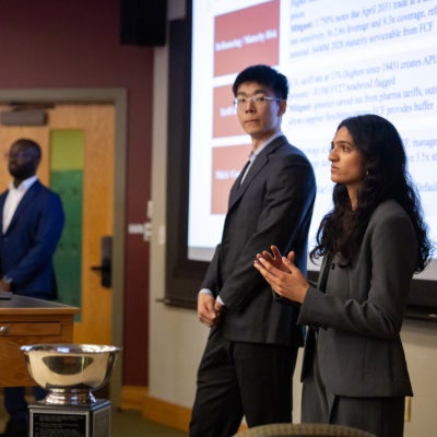Four students in business attire make a presentation in front of a projection screen in a lecture hall.