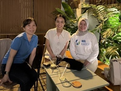 Pera Sihite with two women, smiling with the backdrop of a coffee shop with plants and bamboo fixtures.