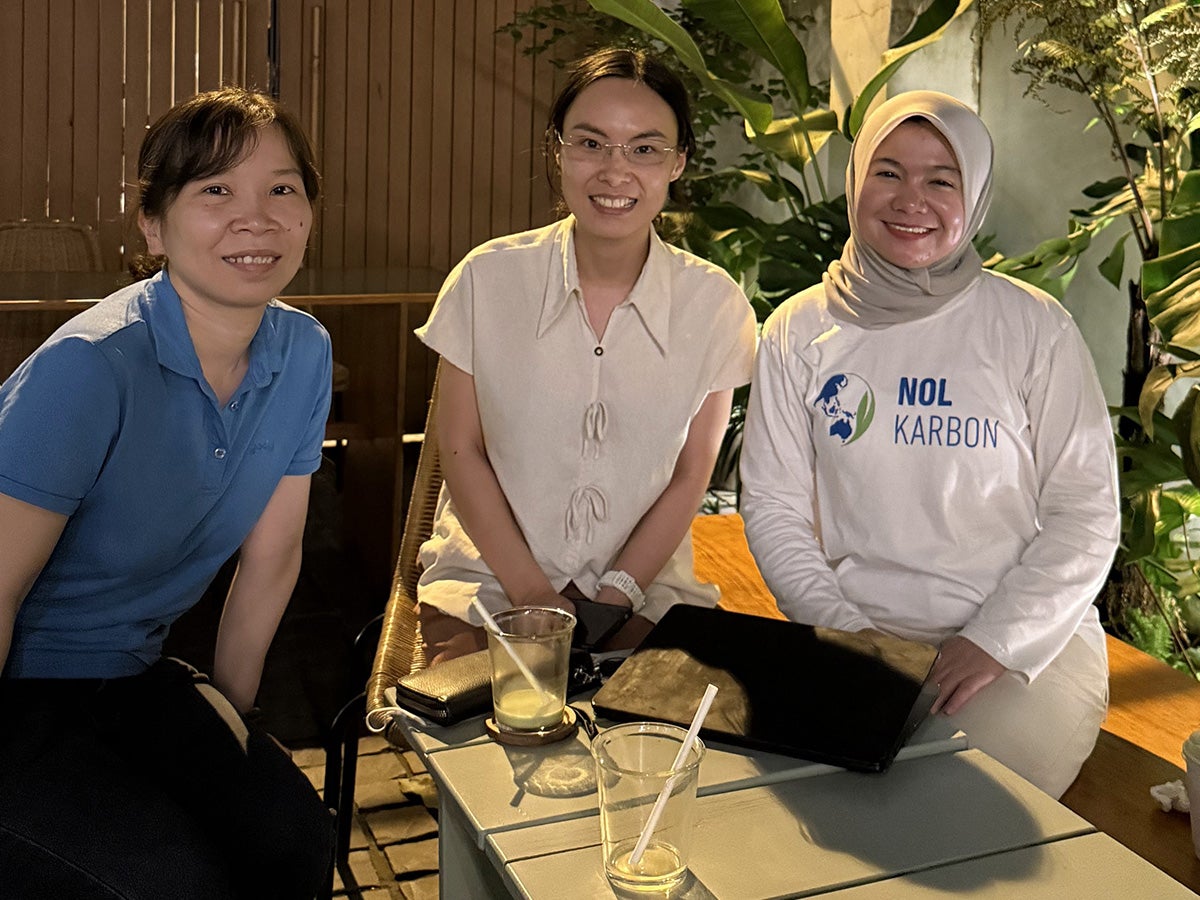 Pera Sihite with two women, smiling with the backdrop of a coffee shop with plants and bamboo fixtures.
