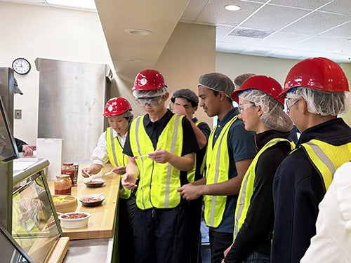 A group of students with hard hats and safety vests tour Giovanni Foods and attend a tasting of sauces.
