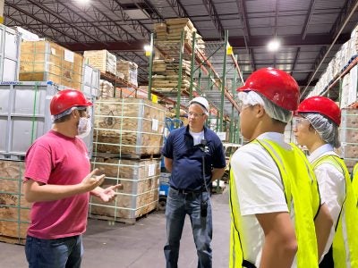 A group of students wearing hard hats speaking with a representative from Giovanni Foods in a warehouse.