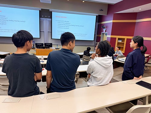 A group of students in a classroom viewing a large screen.