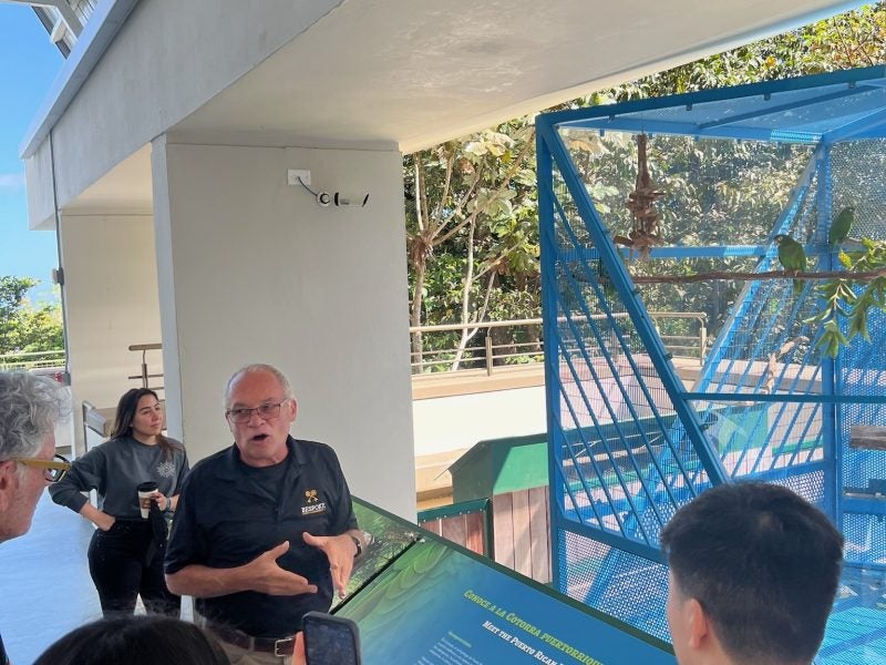 A person speaks in front of birds in a cage.