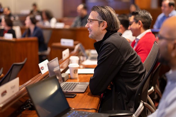 A smiling man sits in a lecture hall with his laptop.