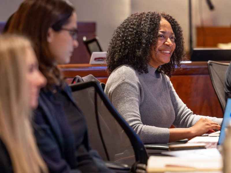 A smiling woman sits in a classroom with her laptop