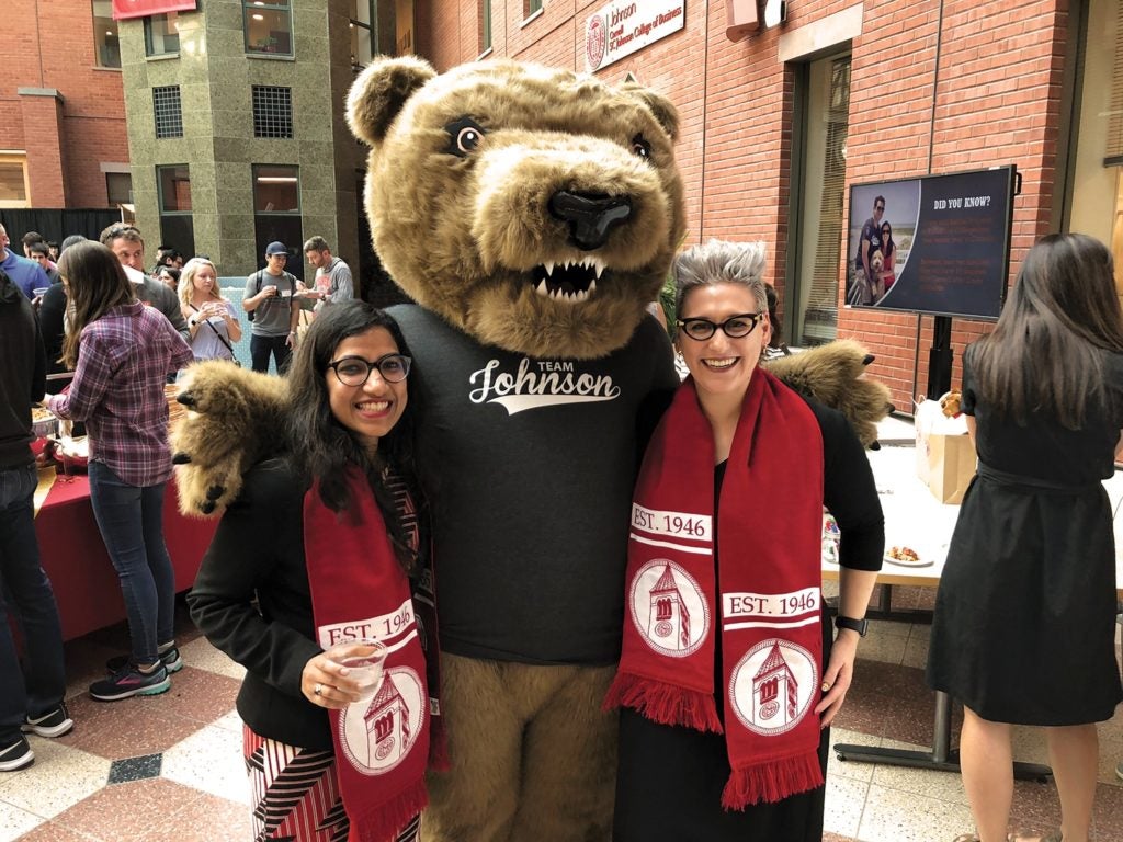 A Cornell bear mascot suited figures stands between two young women Cornell MBA alumnae within the Dyson Atrium at Sage Hall, home of the Cornell MBA at the Johnson School