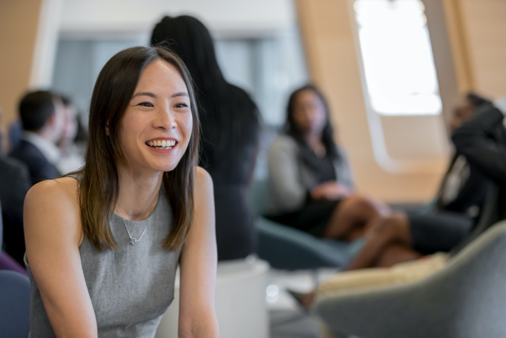 A young woman in business formal attire sits in a sunlit reading room, smiling while speaking to a classmate as part of the Cornell MBA program